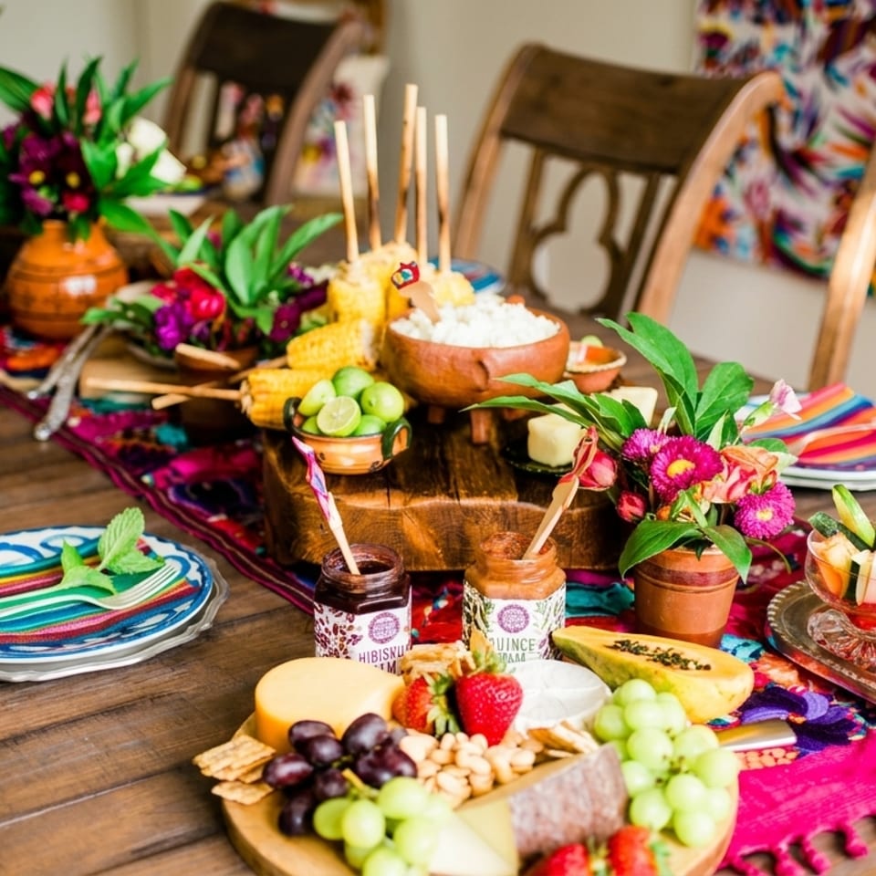 A vibrant, colorful table spread with Mexican food, including elote, fruit, and cheese, with bright decor.