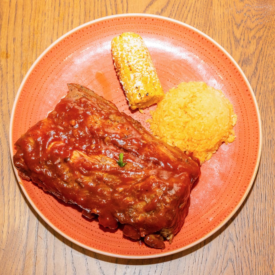 Barbecue-glazed ribs served with yellow rice and grilled corn on a coral plate atop a wooden table.