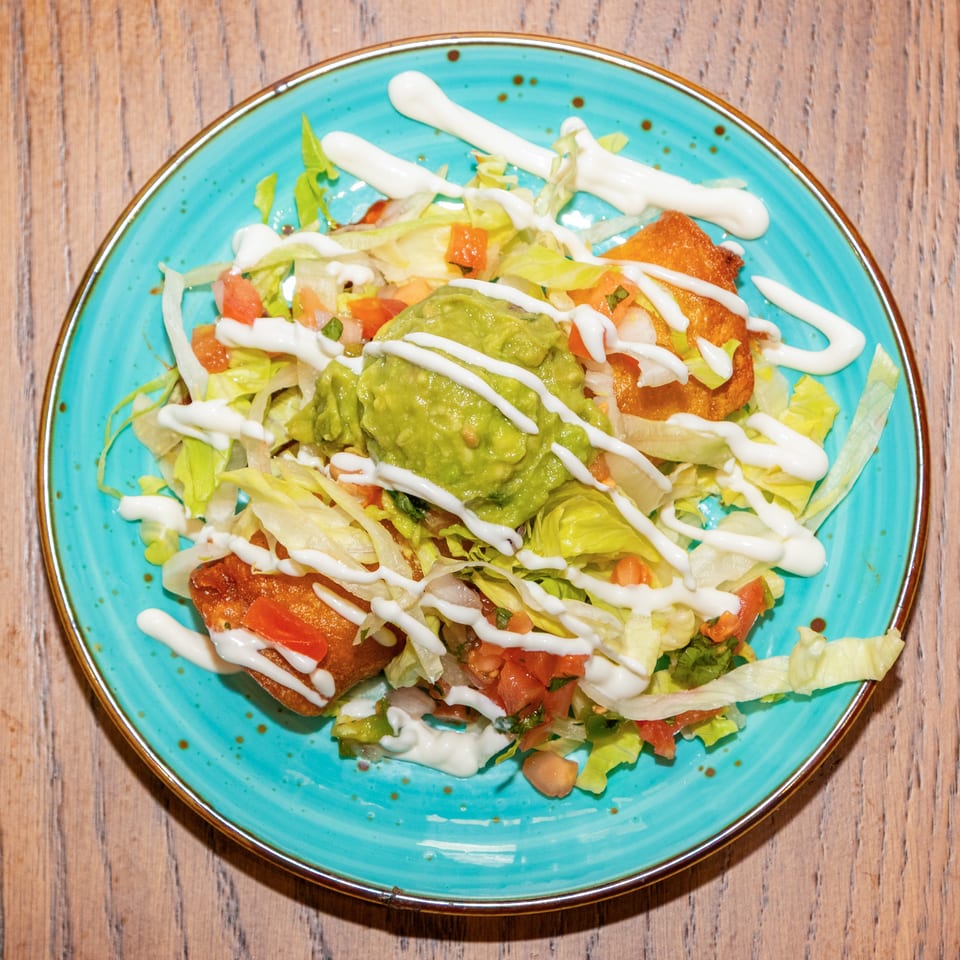 Overhead view of a turquoise plate filled with salad, guacamole, diced tomatoes, crispy tortilla pieces, and drizzled white sauce.